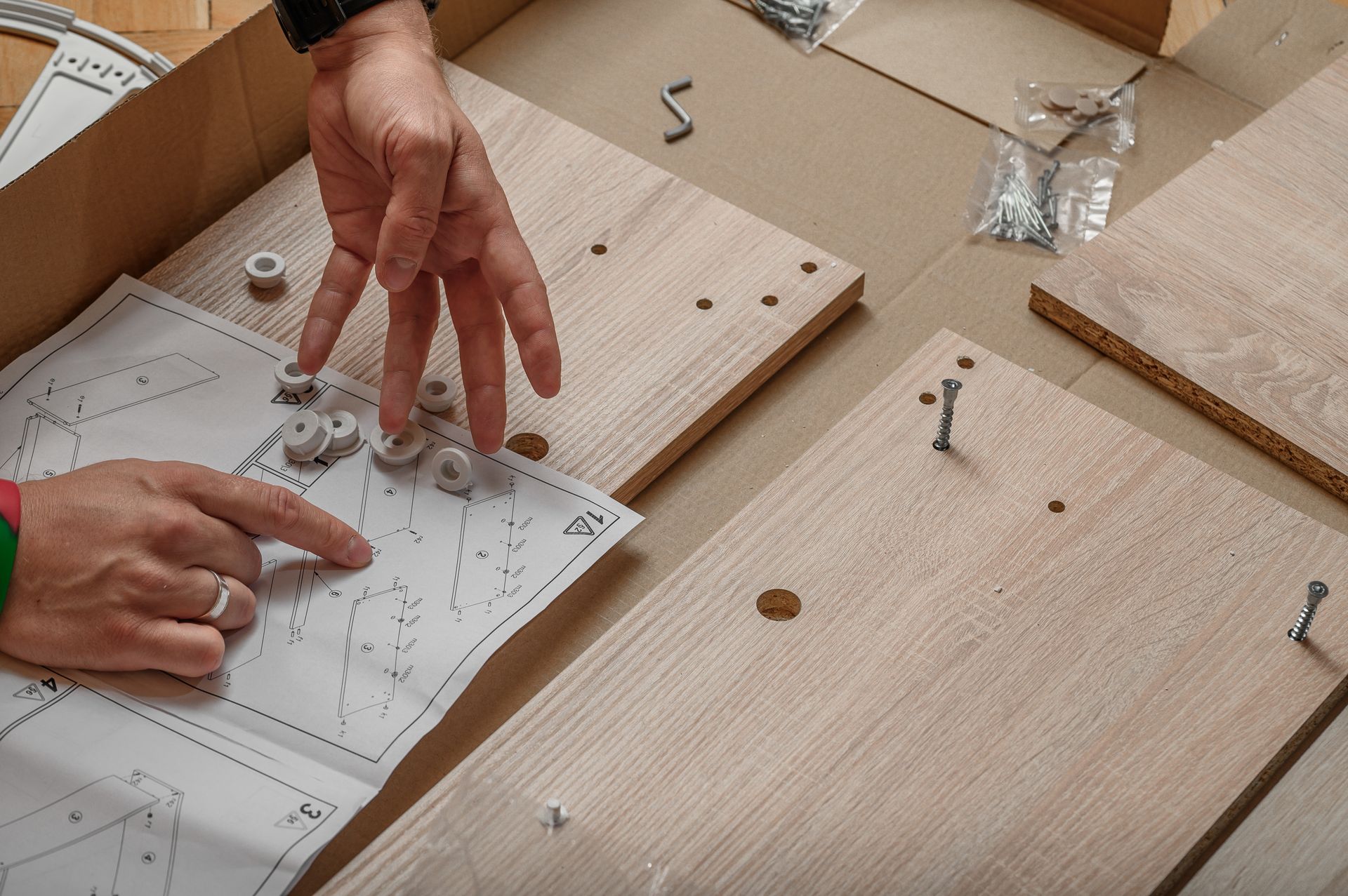 Des mains pointent du doigt les instructions de montage et des panneaux de bois avec la quincaillerie, illustrant un projet de meuble en kit.