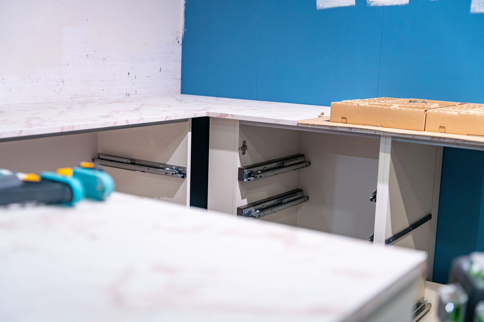Des armoires de cuisine inachevées, avec des glissières de tiroir installées, se détachent sur un mur bleu et des comptoirs clairs.