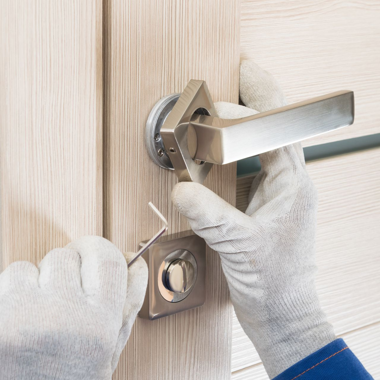 Des mains gantées de blanc utilisent une clé Allen pour serrer une vis sur une poignée de porte moderne en nickel brossé.
