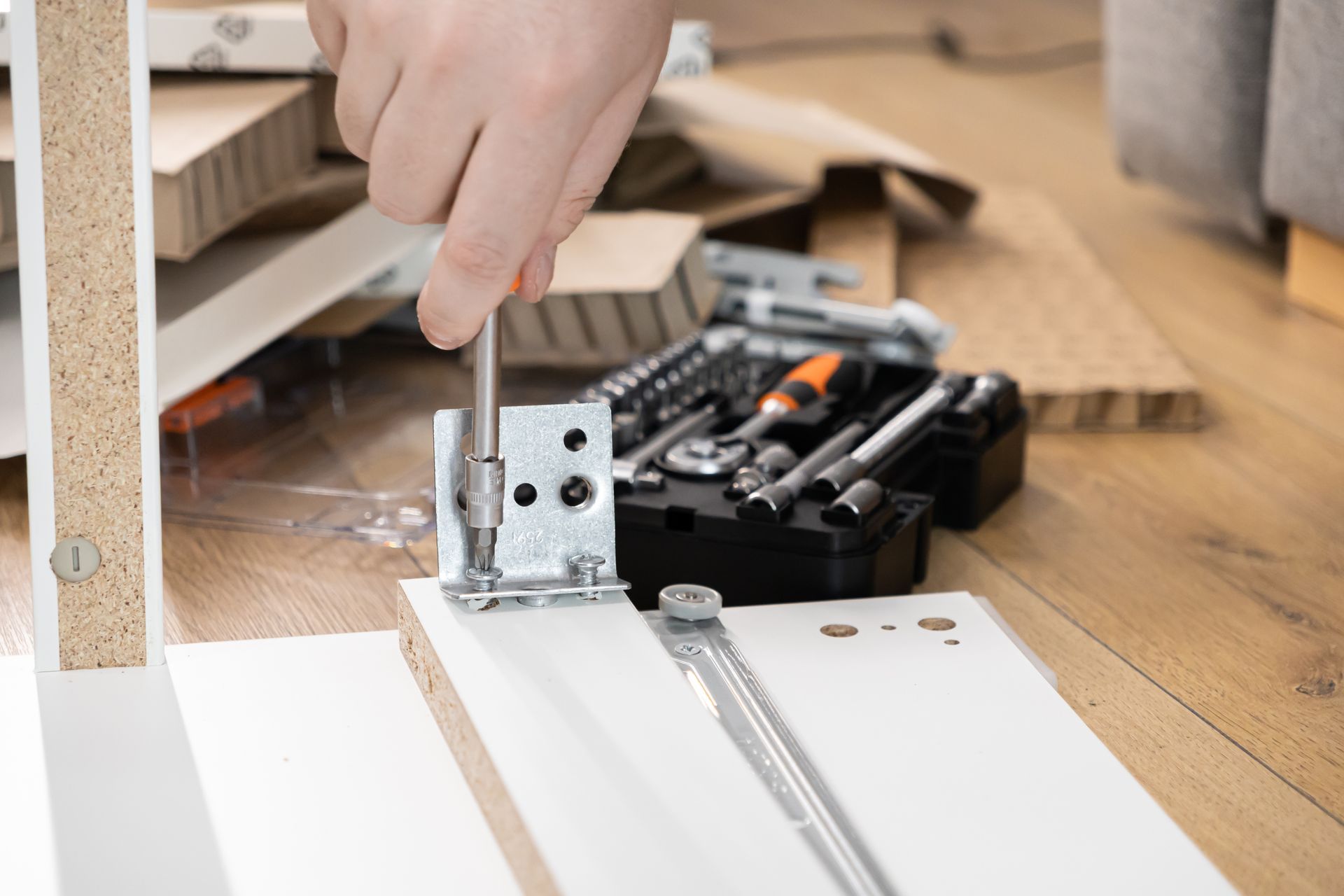 Lors du montage d'un meuble, une personne utilise un tournevis pour fixer une équerre métallique sur un panneau de particules blanc.