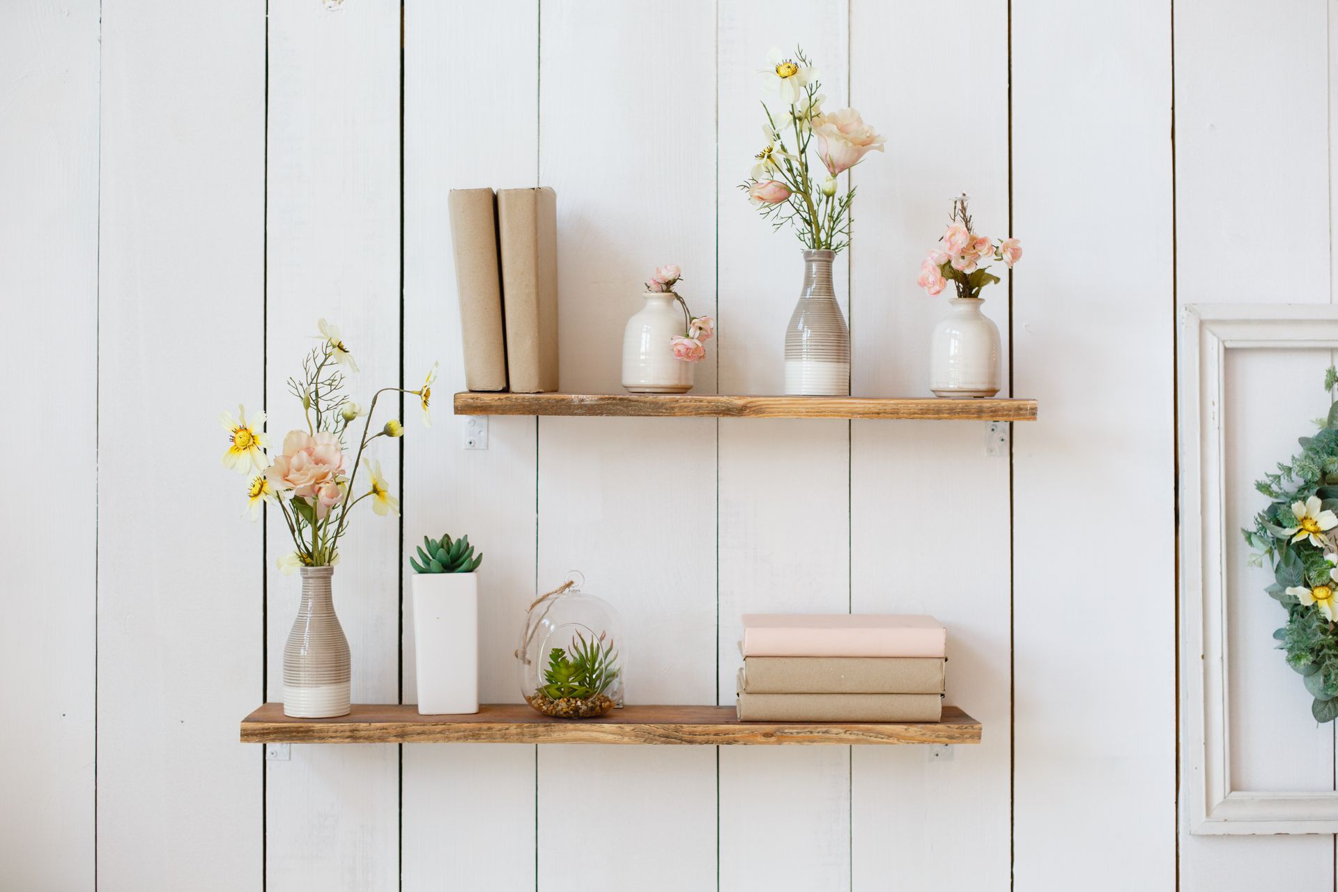 Deux étagères en bois fixées sur un mur lambrissé blanc supportent des vases avec des fleurs, des livres et des petites plantes en pot.