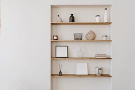 Un mur avec quatre étagères en bois clair sur fond blanc, où sont disposés des vases, des bougies, des livres et des objets décoratifs.