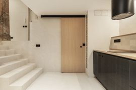 Un intérieur moderne et minimaliste avec une porte coulissante en bois, un escalier blanc et un comptoir de cuisine sombre surmonté d'une hotte ronde.