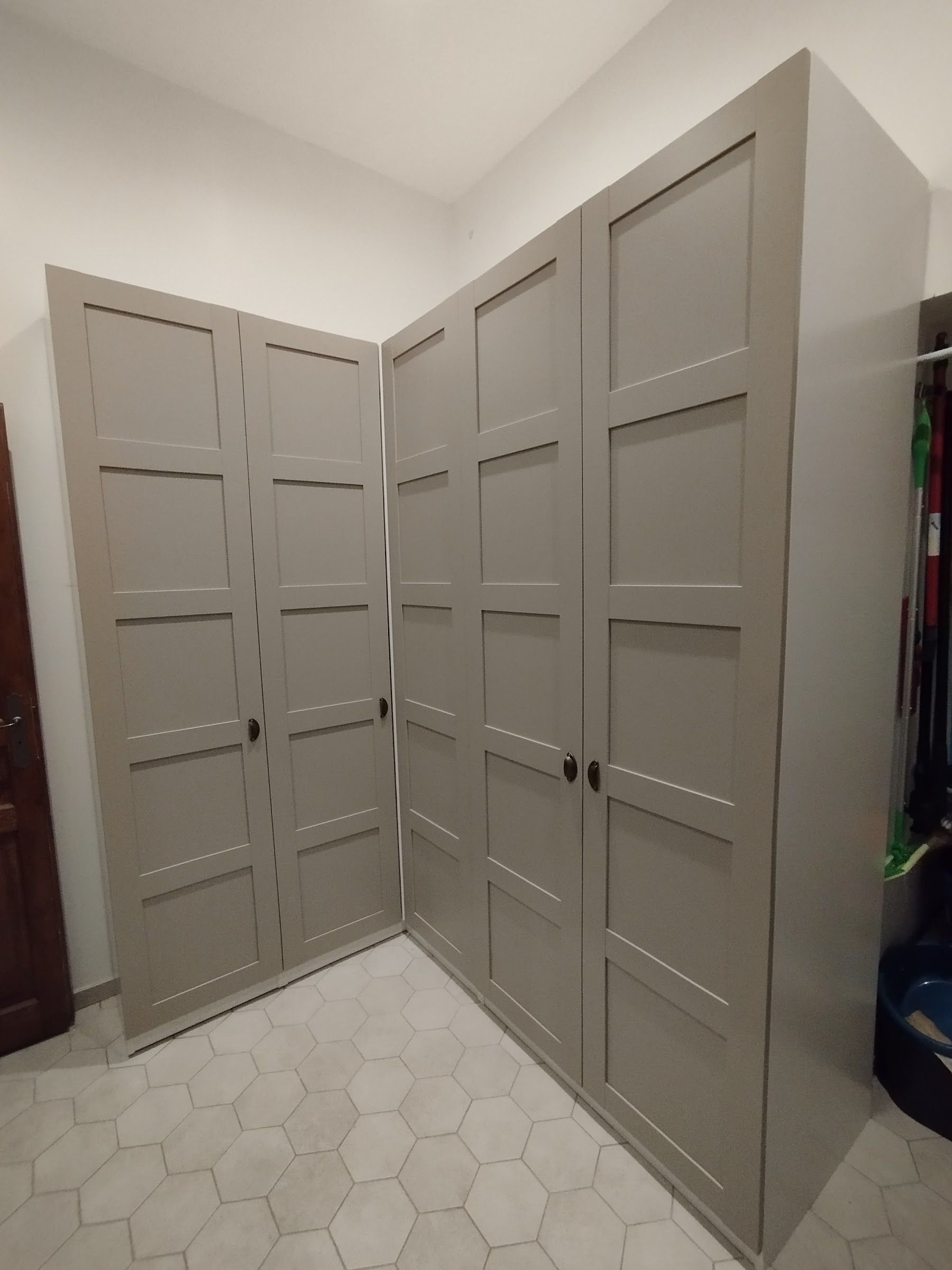 Un coin de pièce orné de hautes armoires en bois gris clair à panneaux, se détachant sur des murs blancs et un sol carrelé.