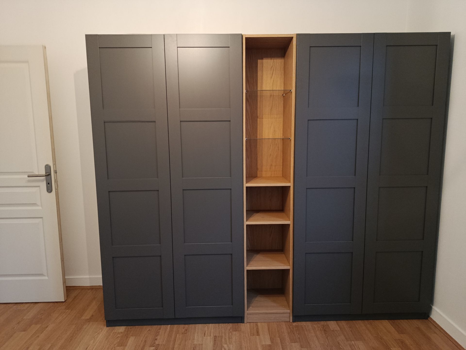 Une grande armoire à quatre portes gris foncé avec une colonne centrale d'étagères ouvertes en bois clair, posée sur un sol en bois.