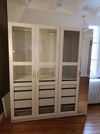 Une armoire blanche à trois compartiments avec portes vitrées, tiroirs intérieurs et panneau latéral miroir, dans une pièce au parquet.