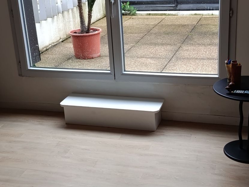 Un petit cache-radiateur blanc et rectangulaire est posé sur un parquet clair, devant une porte-fenêtre coulissante donnant sur une terrasse.