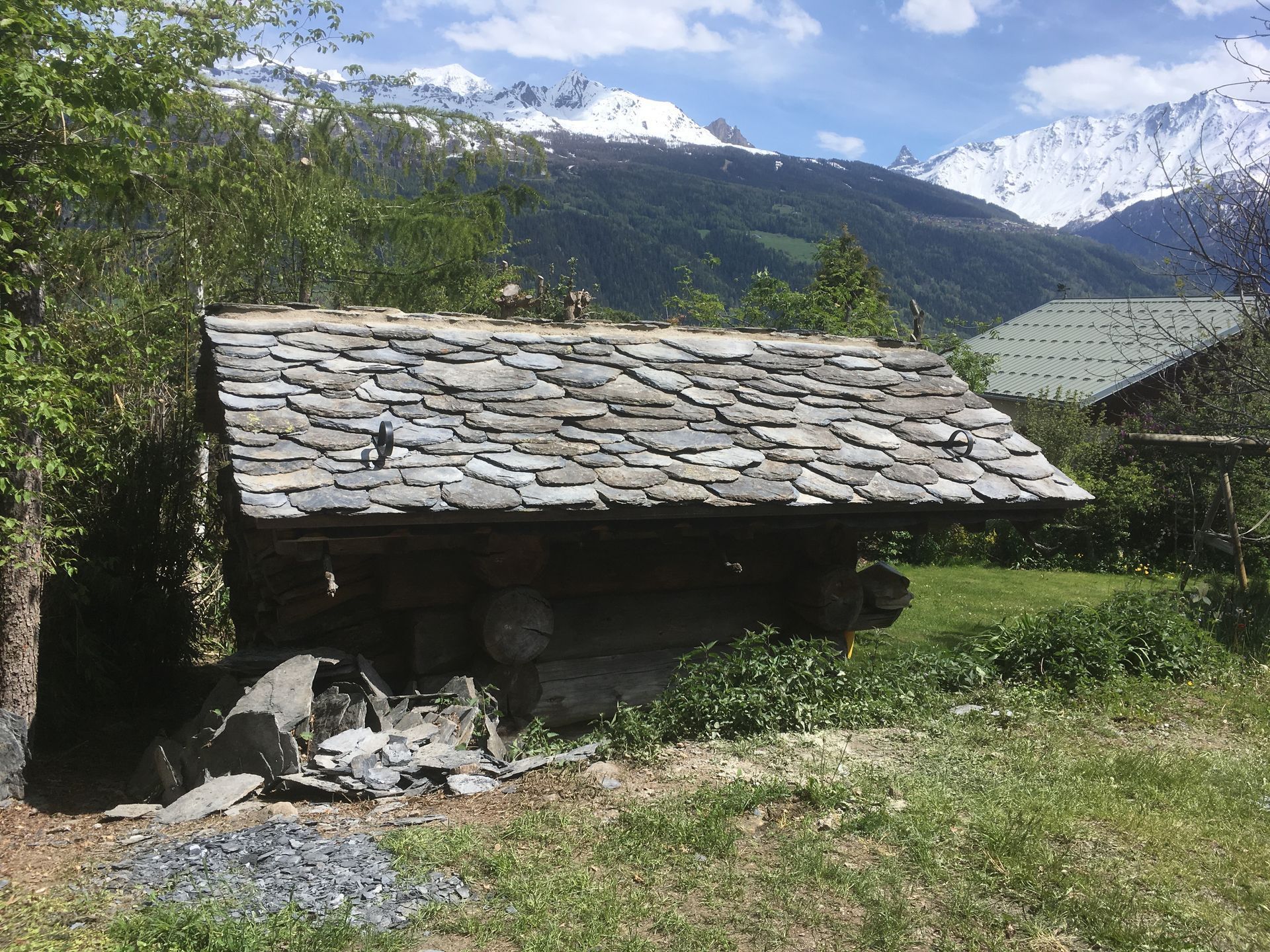 Cabane rustique au toit de pierre dans une vallée montagneuse herbeuse avec des sommets enneigés en arrière-plan