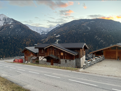 Des chalets en bois bordent une route de montagne, avec des collines saupoudrées de neige au crépuscule.