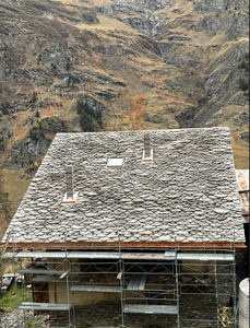 Maison en construction au toit de pierre, avec une montagne rocheuse en arrière-plan.