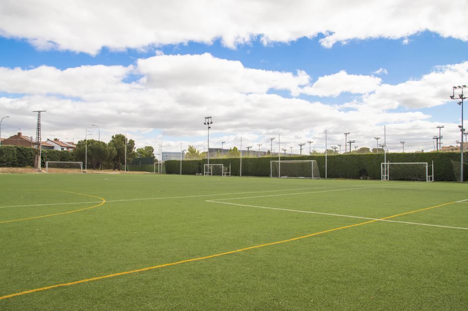 Campo de fútbol verde con porterías blancas, bajo un cielo azul con nubes esponjosas.