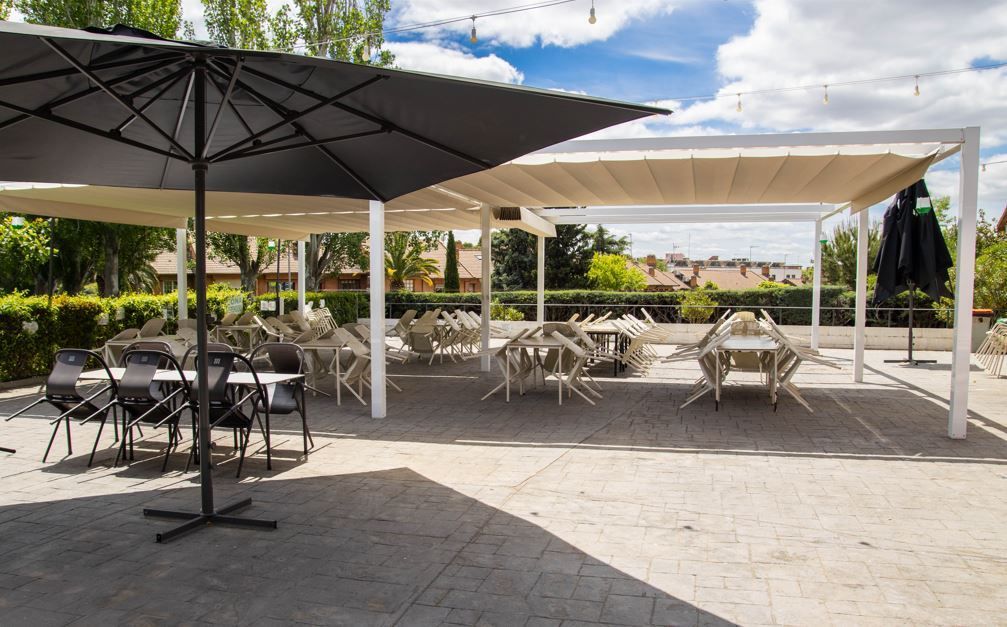 Comedor al aire libre con mesas y sillas bajo techo retráctil y sombrilla en un día soleado.