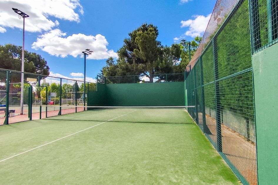 Pista de pádel verde con red, rodeada por una valla verde, bajo un cielo azul con nubes.