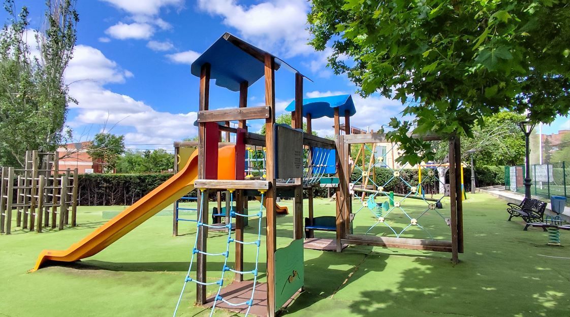 Zona de juegos con estructura de madera, tobogán y césped verde bajo un cielo azul.