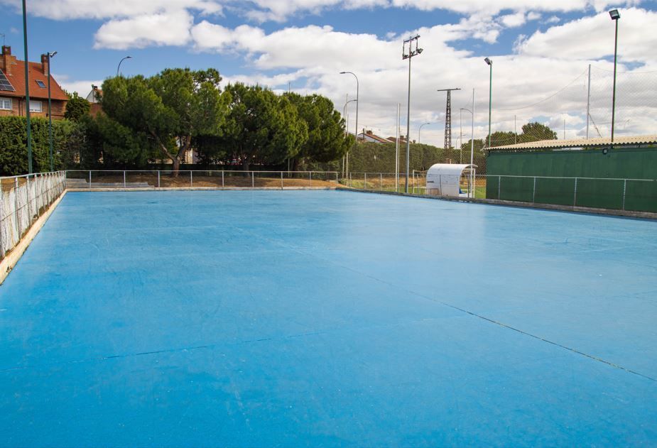 Pista deportiva exterior azul con valla y árboles alrededor bajo un cielo nublado.