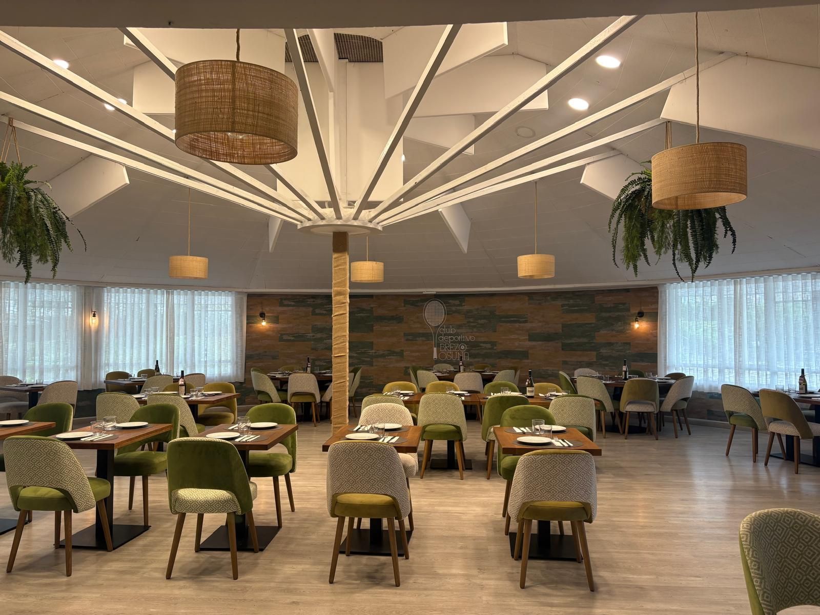 Interior de restaurante con mesas y sillas, techo decorativo y plantas colgantes; combinación de colores verde y beige.