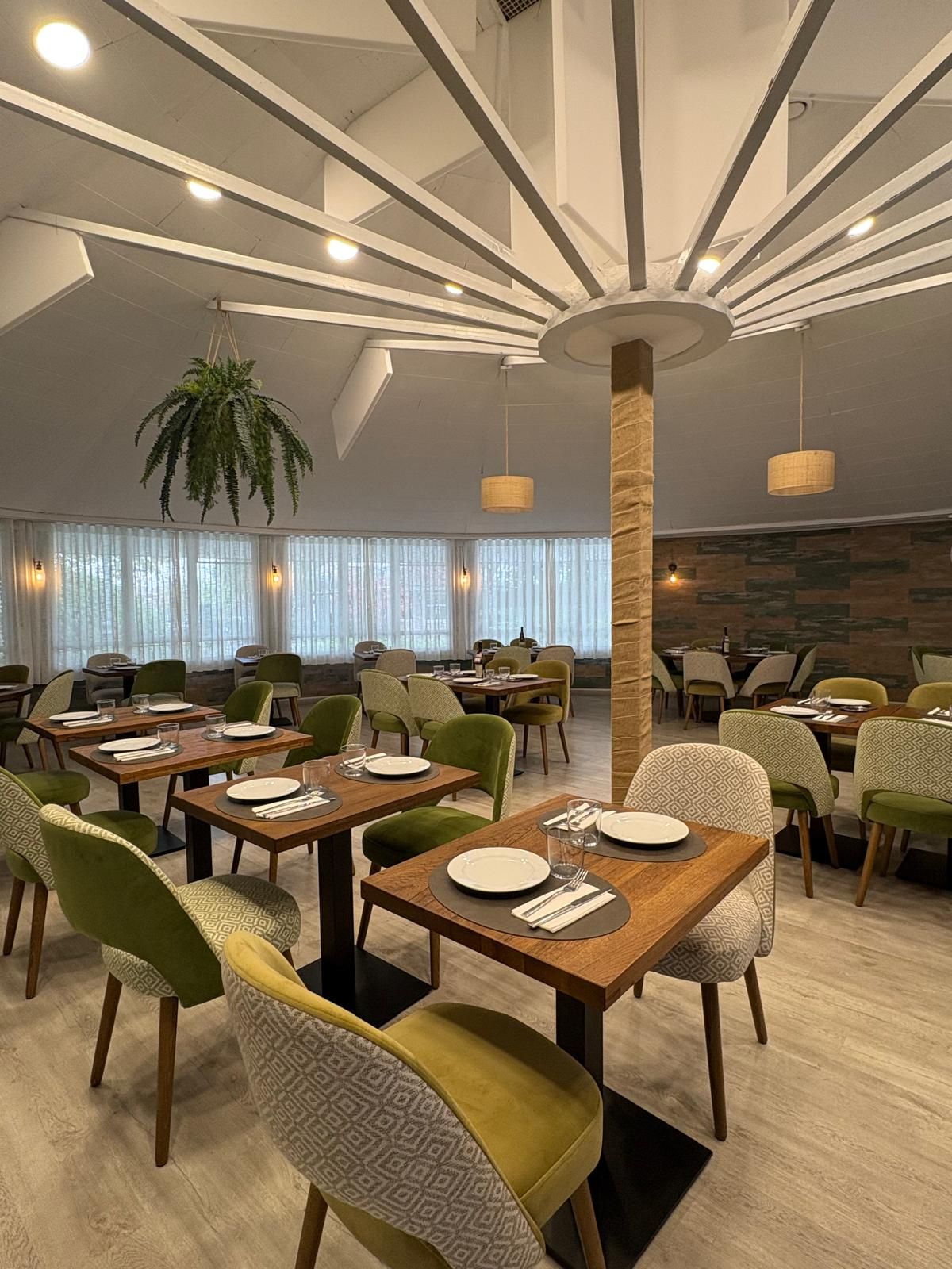 Interior del restaurante con mesas, sillas y plantas colgantes. Paredes y techo de color claro con vigas decorativas.