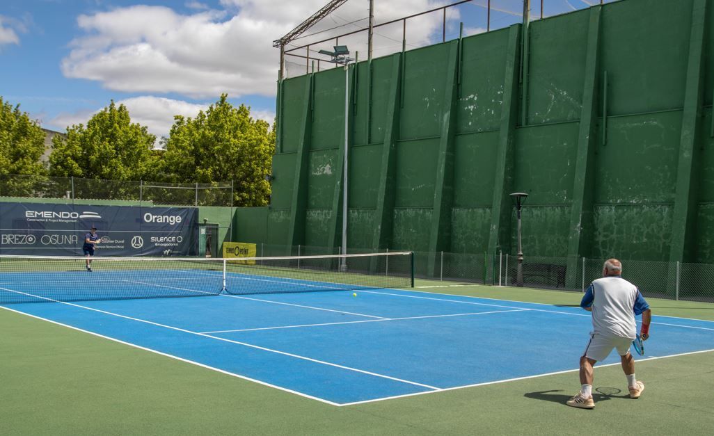 Dos hombres jugando al pádel en una pista azul, con una valla verde al fondo, en un día soleado.