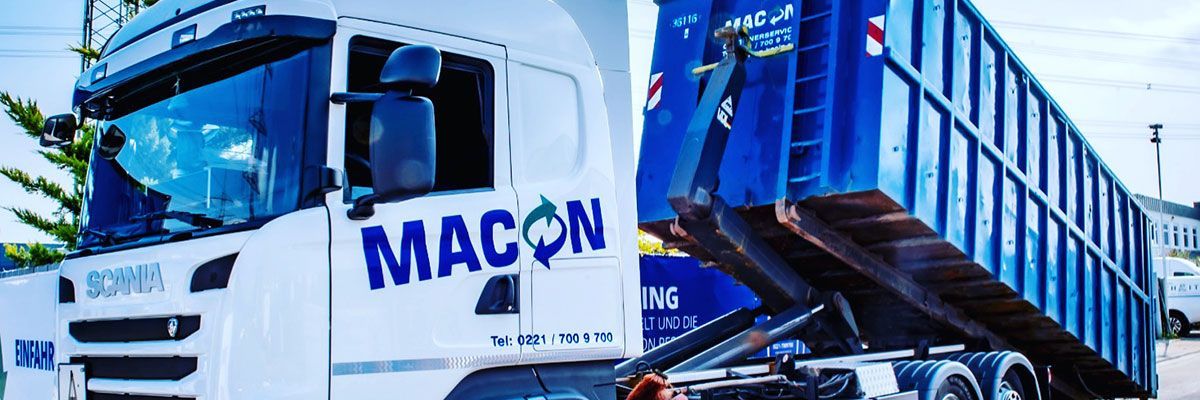 Weißer Lastwagen mit der Aufschrift „MACON“ an der Seite, der einen blauen Müllcontainer zieht.