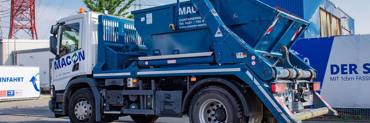 Blau-weißer MACON-Müllwagen mit erhöhtem Container vor einer weißen Wand mit blauen Akzenten.
