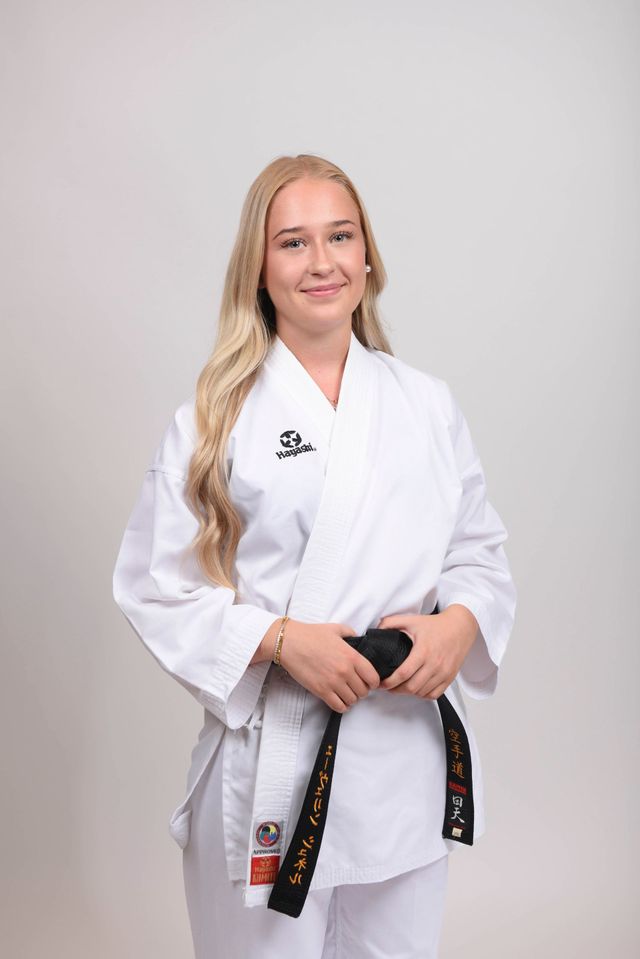 Frau in weißer Karateuniform, schwarzer Gürtel, steht lächelnd vor einem weißen Hintergrund.