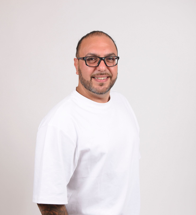 Mann mit Brille und weißem T-Shirt lächelt vor weißem Hintergrund in die Kamera.