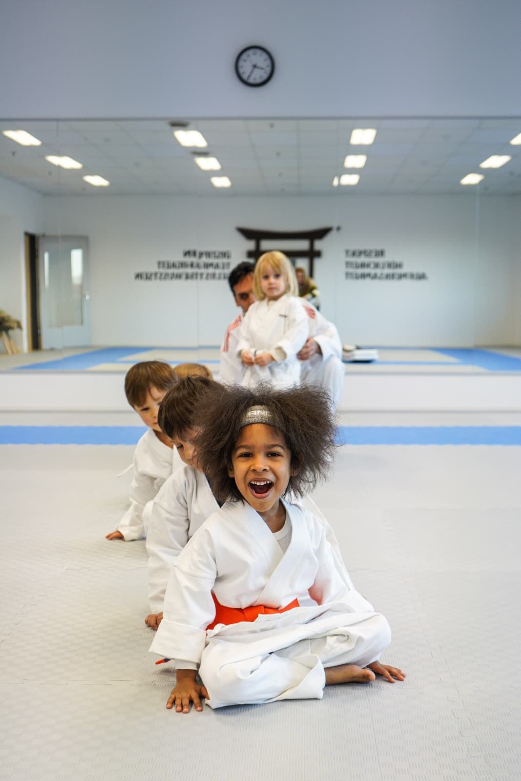 Mädchen in Karateuniform lächelt, posiert mit erhobenen Fäusten, gelber Hintergrund.
