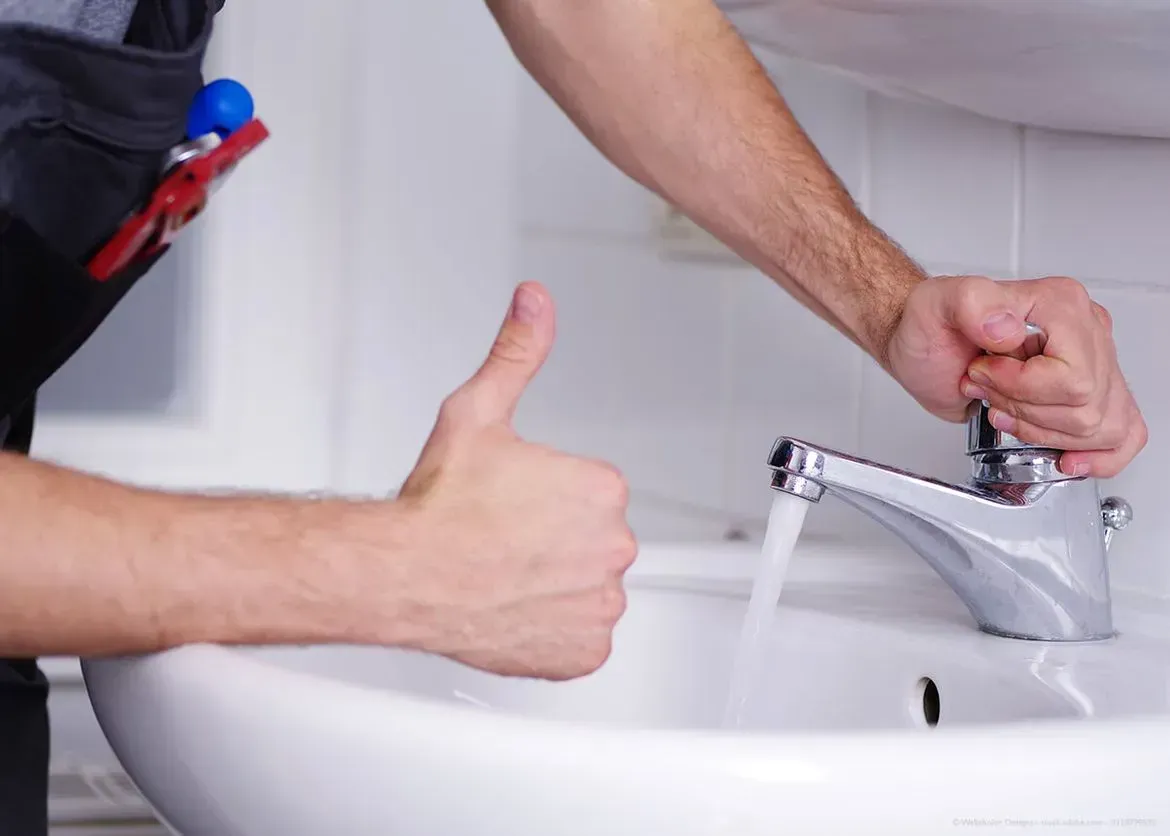 Ein Mann repariert einen Wasserhahn in einem Badezimmer und streckt den Daumen nach oben aus.