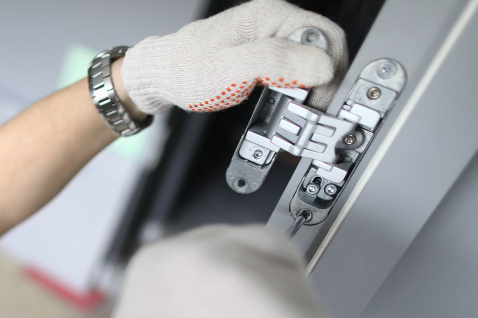 Une personne portant des gants installe une charnière métallique sur un cadre gris.
