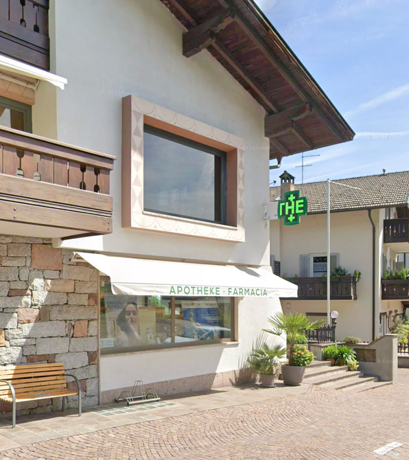 Farmacista in camice bianco sorride, braccia incrociate, in una farmacia con scaffali pieni di medicinali.