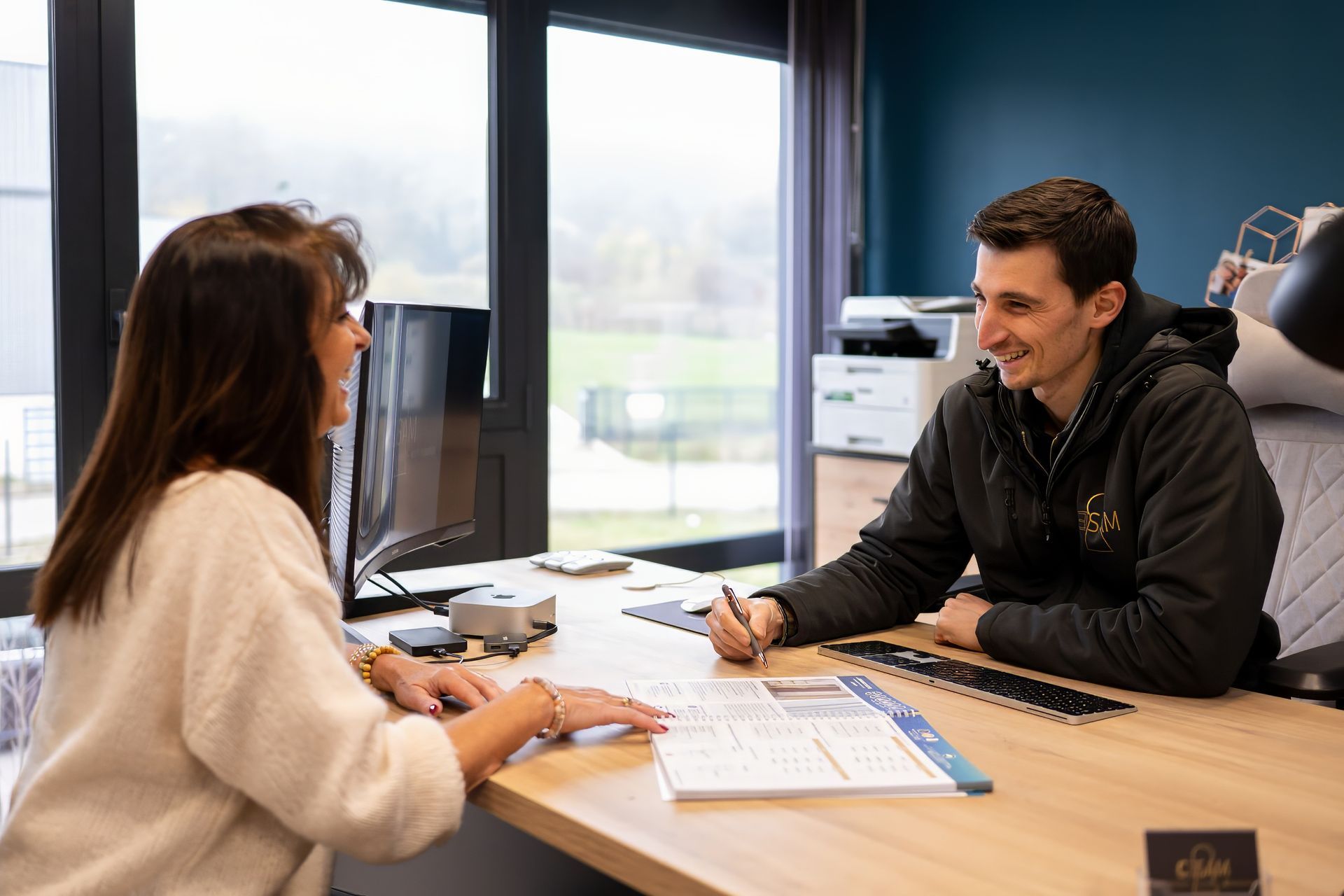 M. Cardon et une femme sont assis à un bureau, des catalogue de menuiserie sont posés entre eux