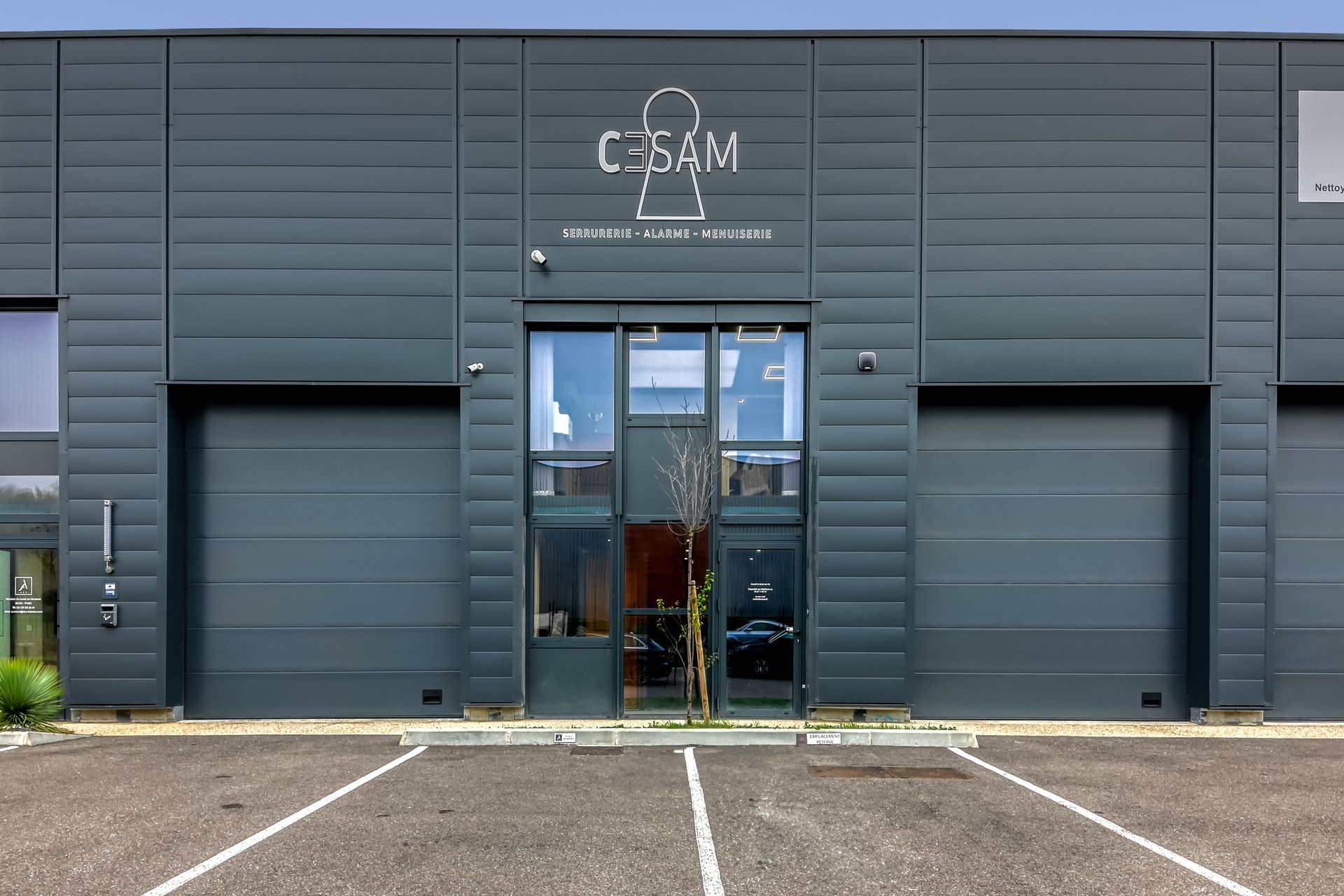 Bâtiment gris foncé avec le logo « CESAM ». Grandes portes de garage, porte centrale.