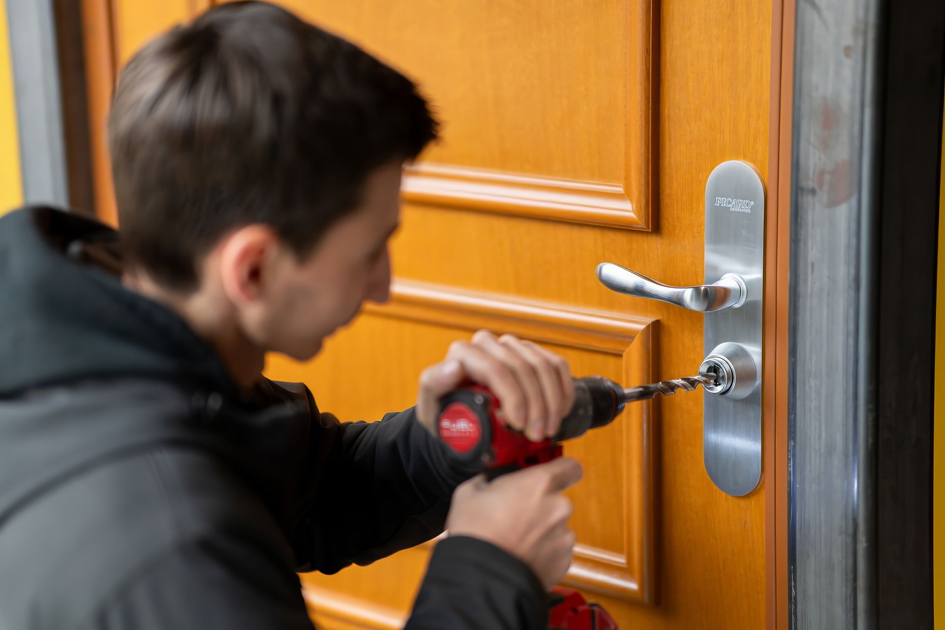 Une personne perce une serrure sur une porte en bois. Elle utilise une perceuse rouge. La porte est orange et argentée.
