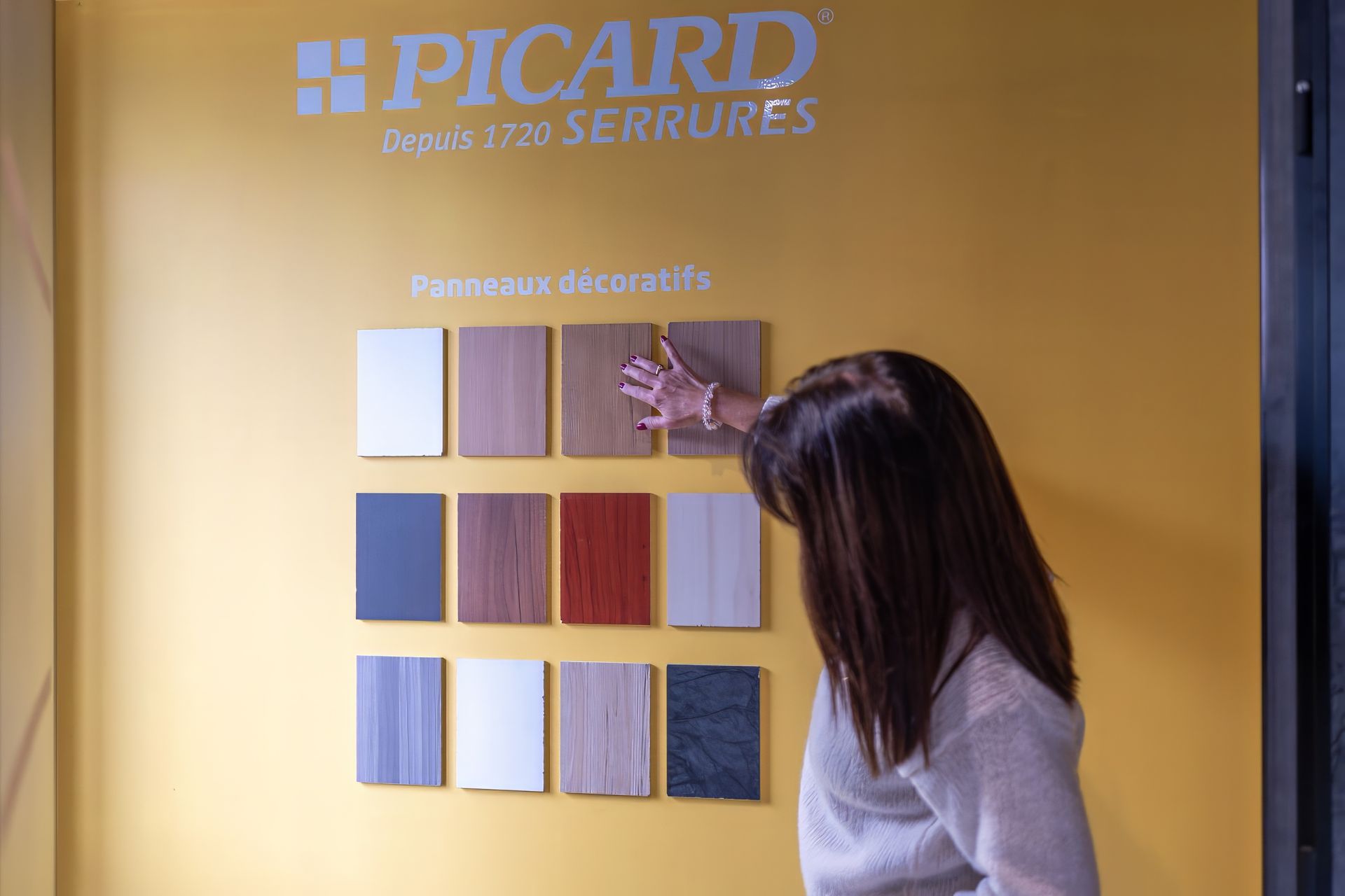 Une femme examine des échantillons de bois sur un mur jaune portant le logo et l'inscription « PICARD ».