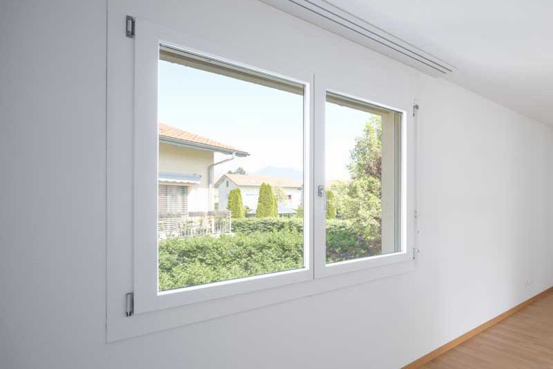 Fenêtre blanche à double vitrage dans une pièce blanche, donnant sur une vue ensoleillée d'arbres et d'une maison.