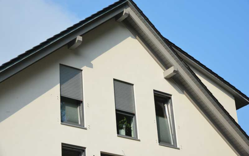 Bâtiment blanc à l'extérieur, avec trois fenêtres munies chacune de stores gris, sous un toit sombre.