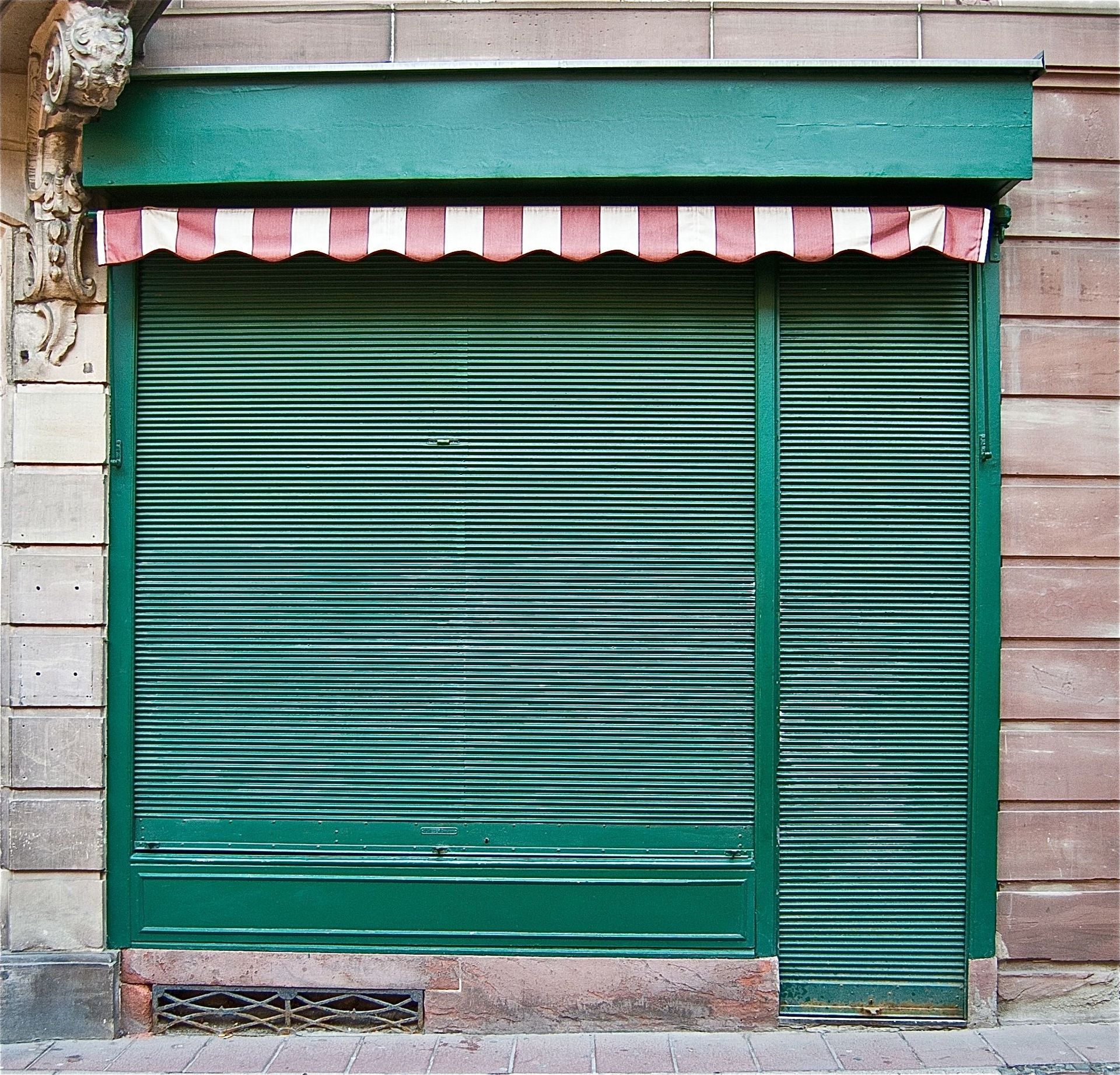 Façade verte avec volets métalliques fermés, auvent à rayures rouges et blanches.