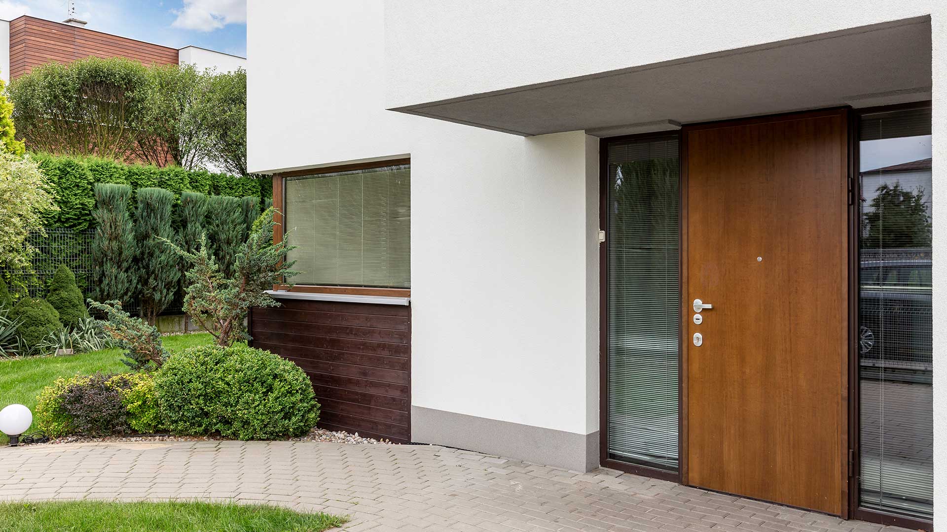 Entrée d'une maison moderne avec porte marron, panneaux de verre et petit jardin.