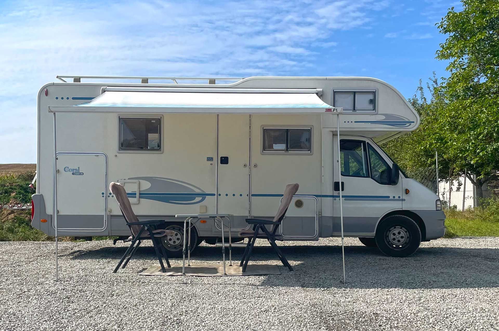 Ein weißes Wohnmobil steht mit einem Tisch und Stühlen auf einem Schotterplatz.