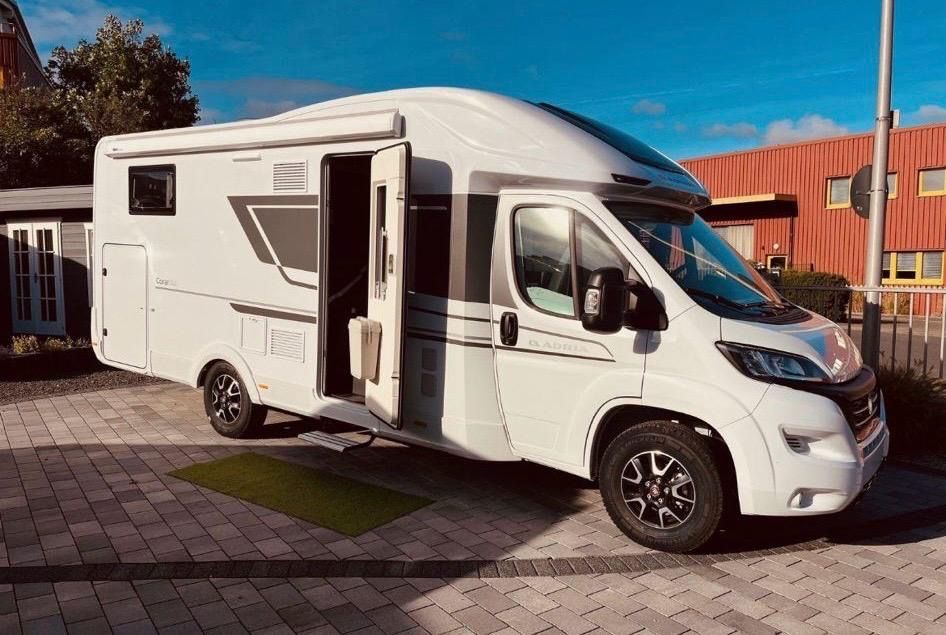 Ein weißes Wohnmobil, Marke Adria Coral Axess 670 SL, steht auf einer gepflasterten Auffahrt vor einem Gebäude.
