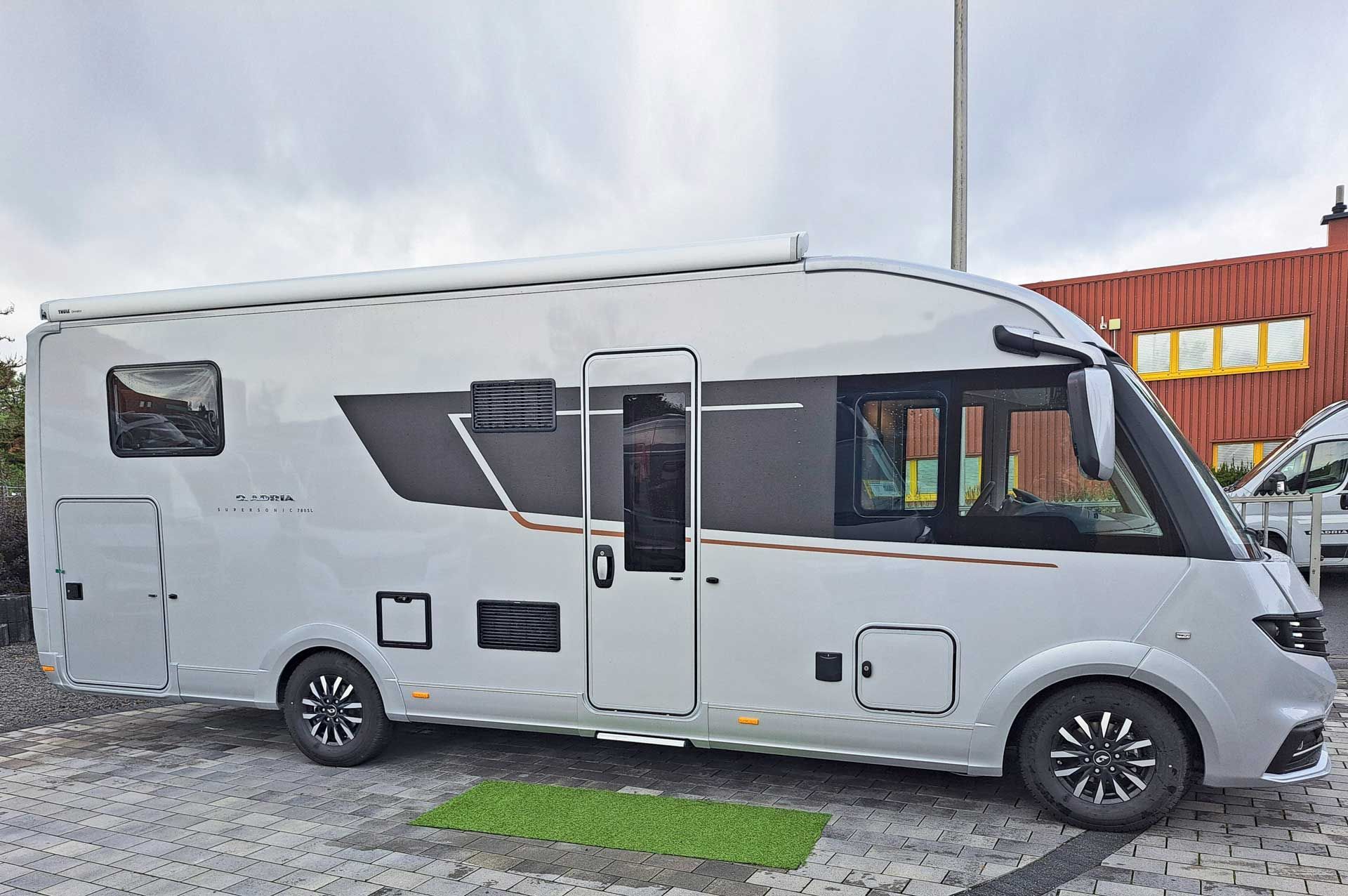 Ein weißes Wohnmobil, Modell Adria Supersonic 780 SL, steht auf einem Parkplatz vor einem Gebäude.