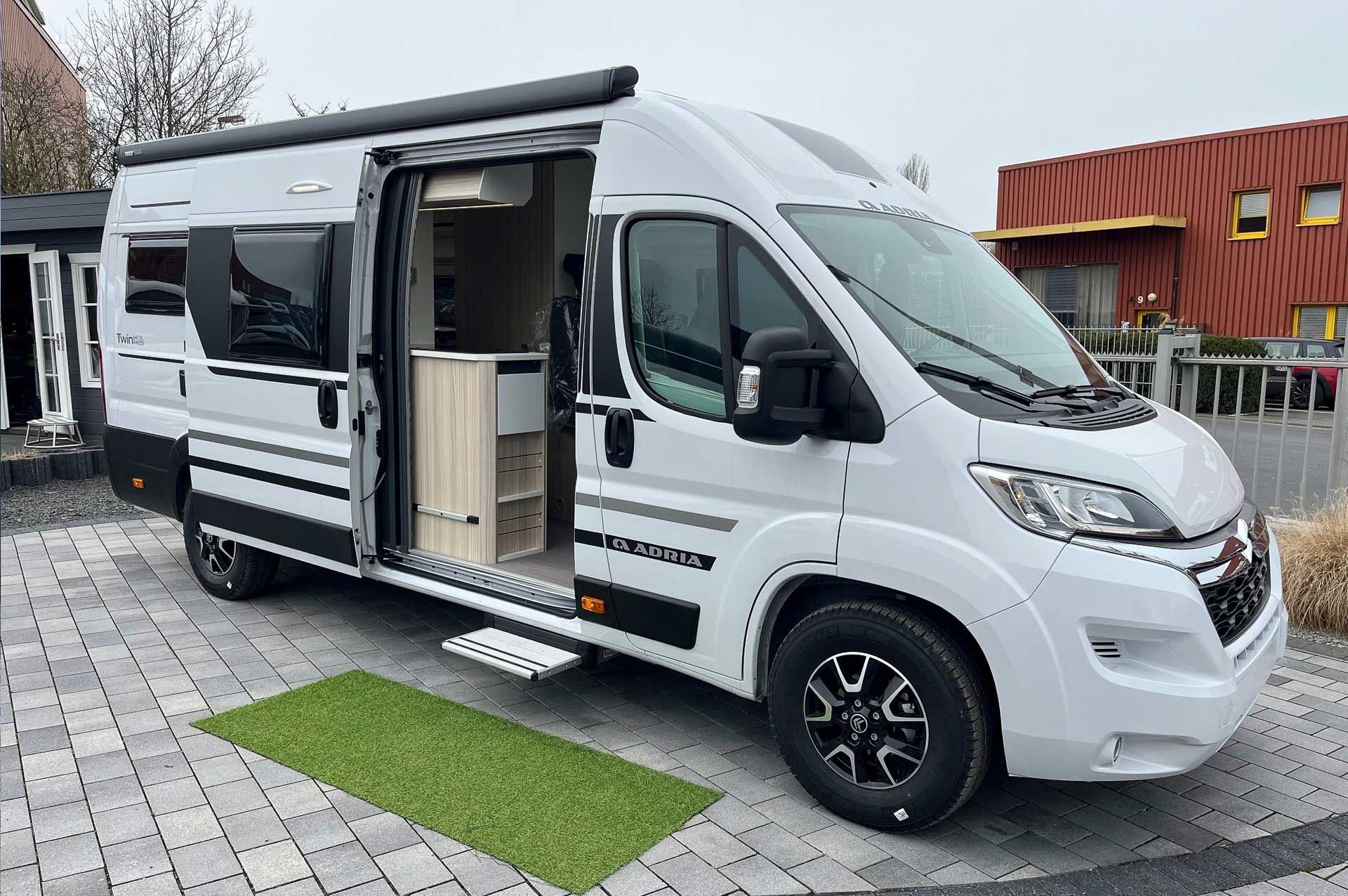 Auf einer gepflasterten Auffahrt steht ein weißes Wohnmobil, Modell Adria Twin ALL-IN 640 SL, mit offener Tür.