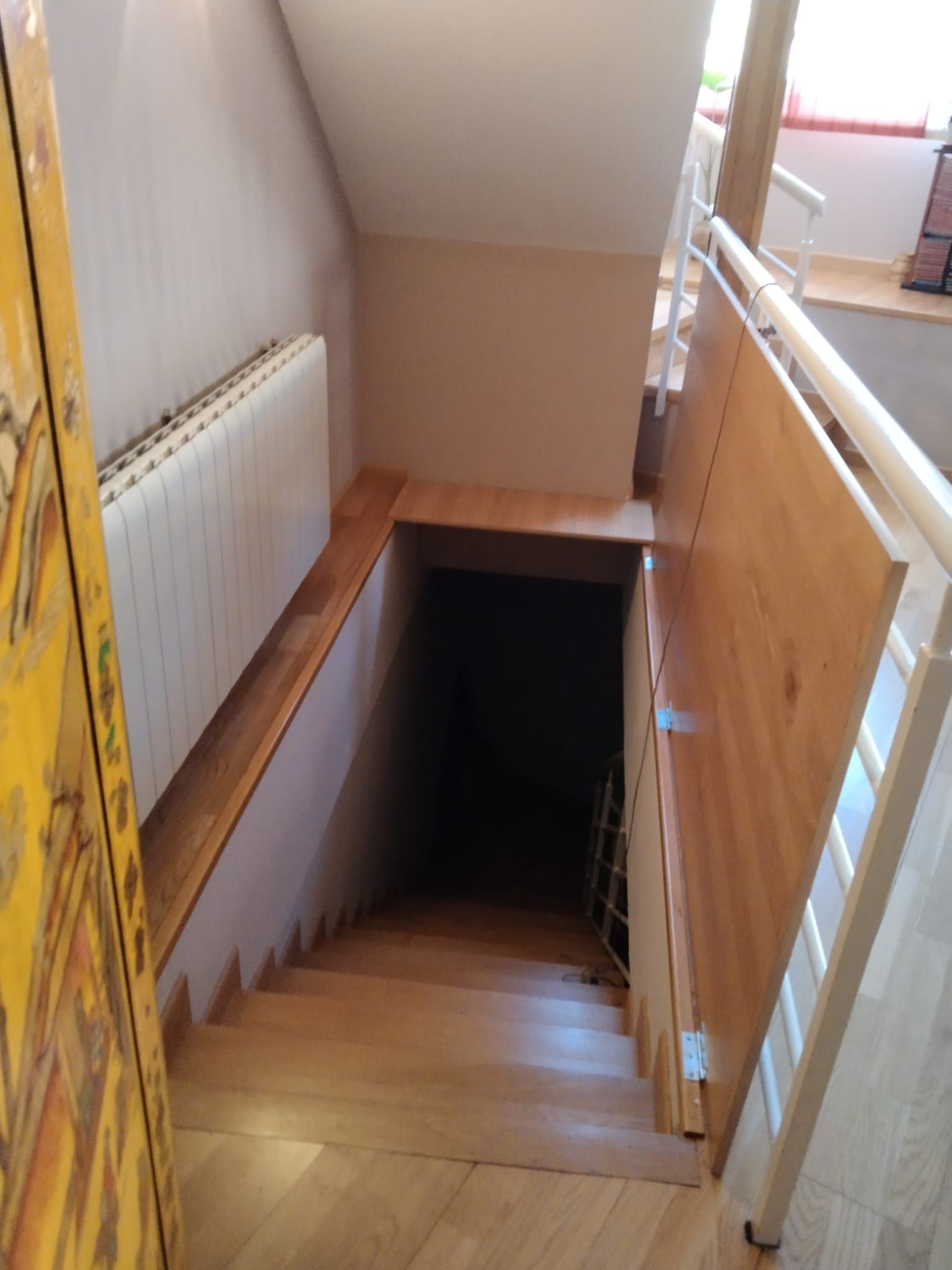 Escalera que desciende hacia la oscuridad. Peldaños y barandilla de madera, radiador blanco, paredes beige y una puerta