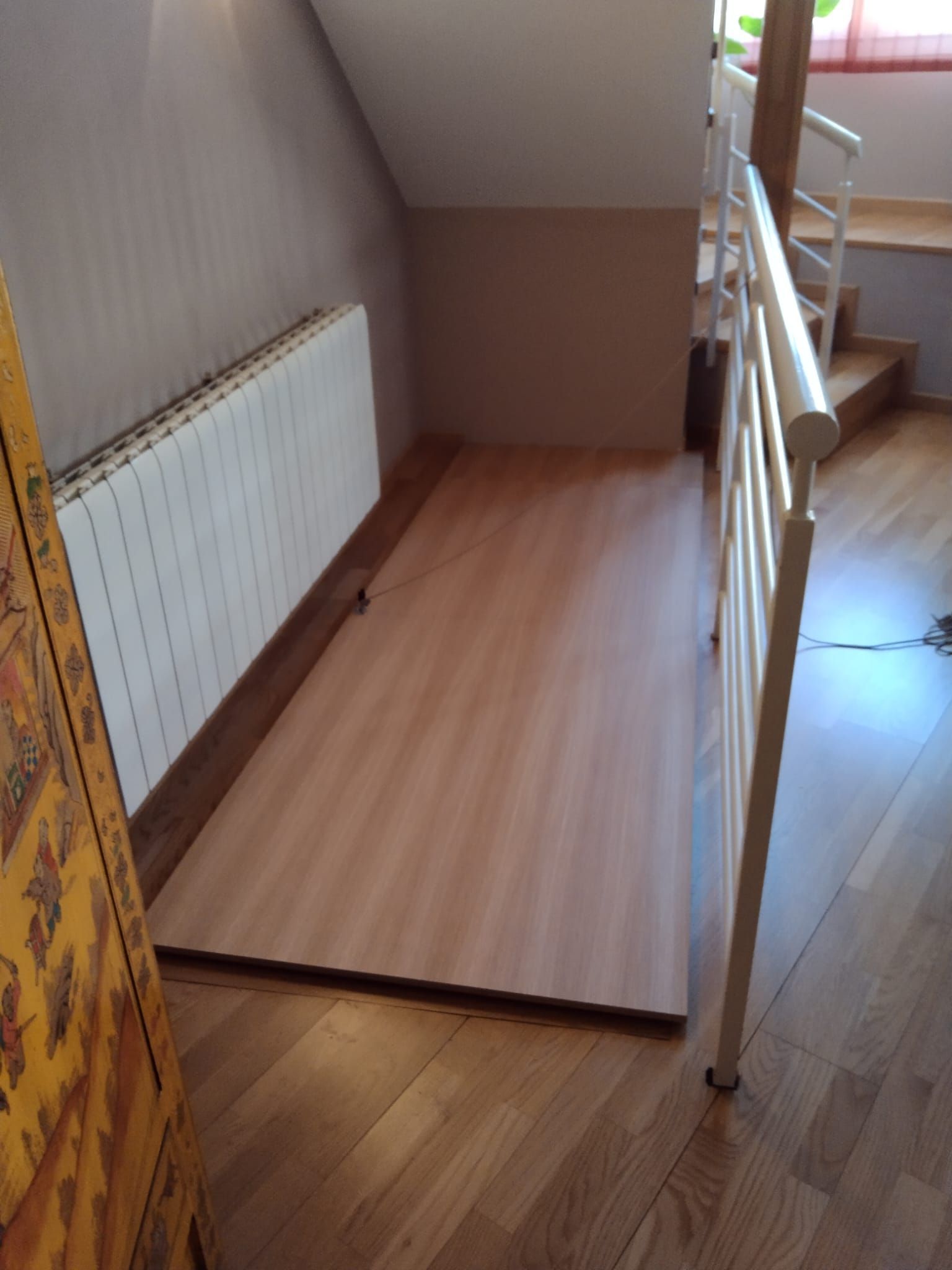 Espacio de suelo de madera junto a un radiador, con barandilla blanca y escaleras al fondo.