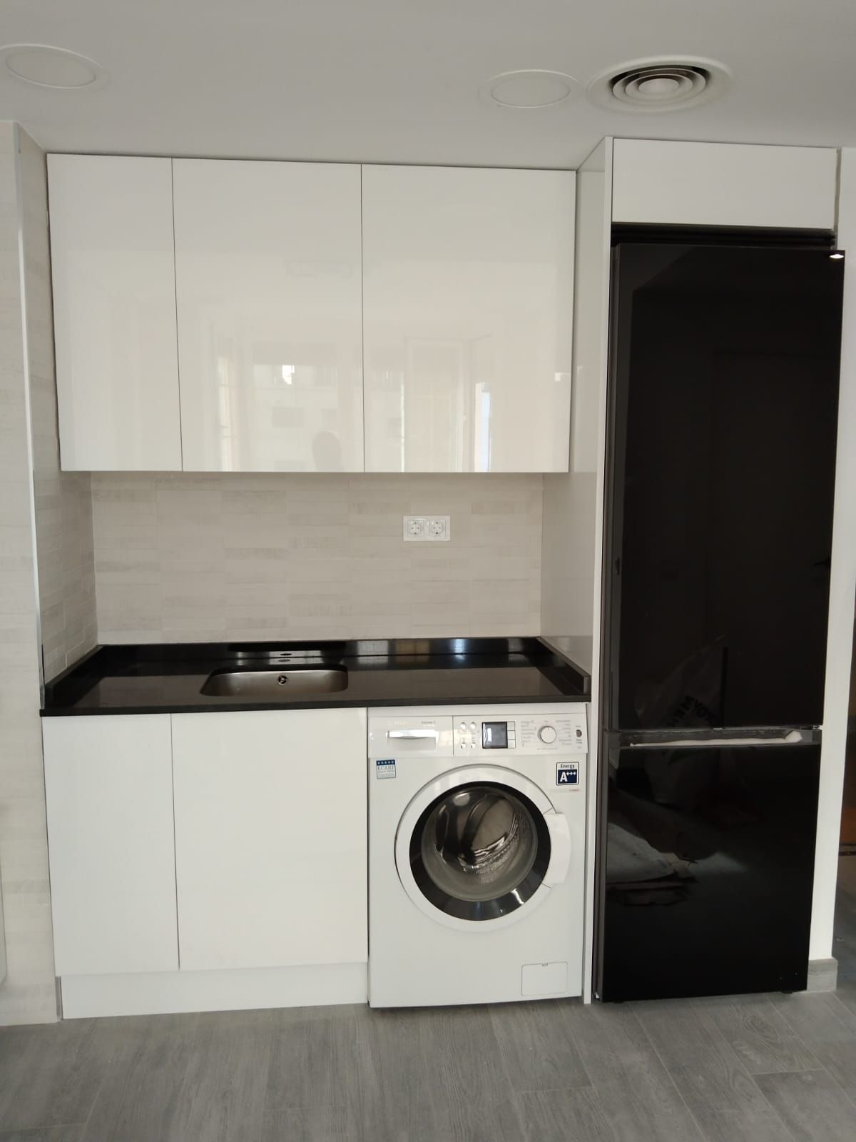 Muebles de cocina blancos, lavadora, encimera y refrigerador negros. Suelo gris y salpicadero beige.