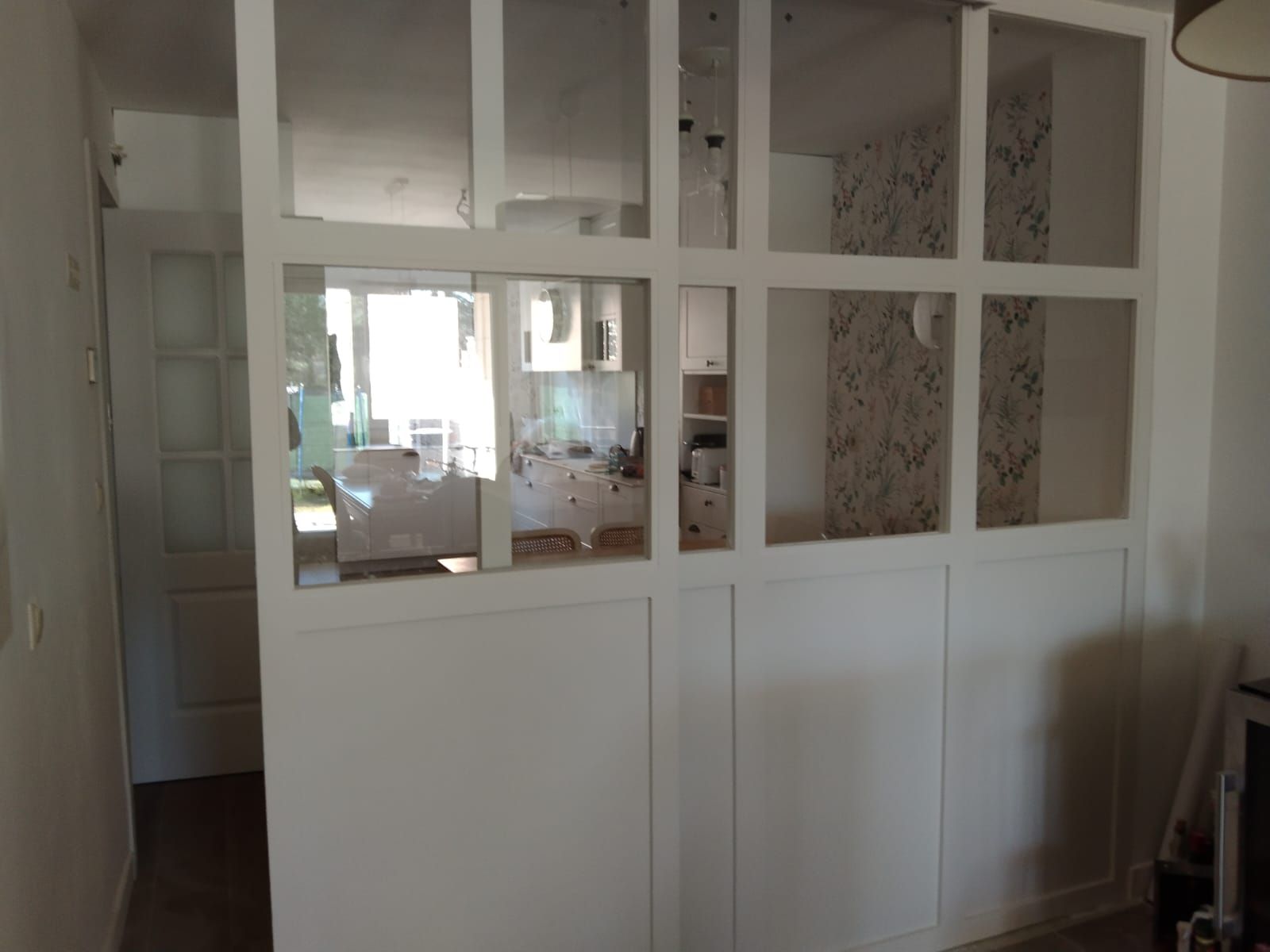 Biombo con marco blanco, paneles de vidrio y paneles inferiores que separan espacios y dejan ver una cocina y un pasillo