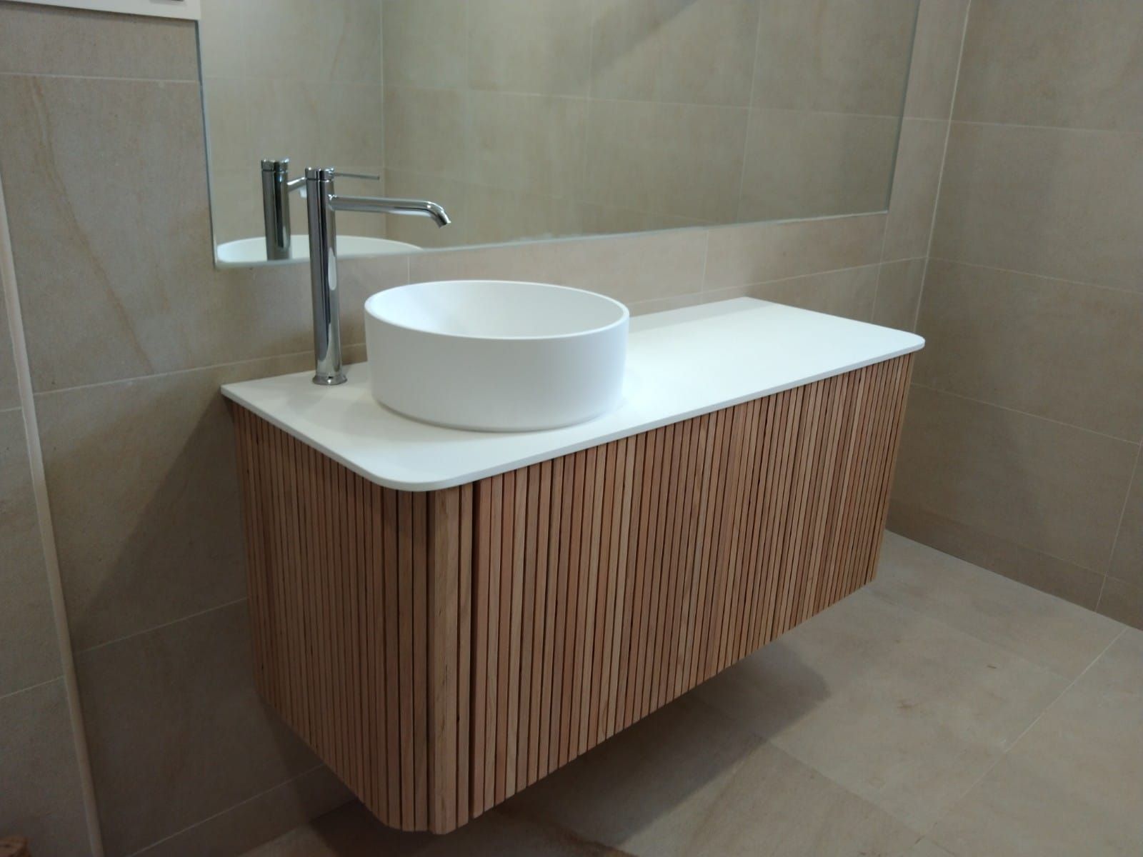 Tocador flotante de madera con encimera blanca y lavabo redondo; grifería cromada; paredes revestidas de azulejos beige.