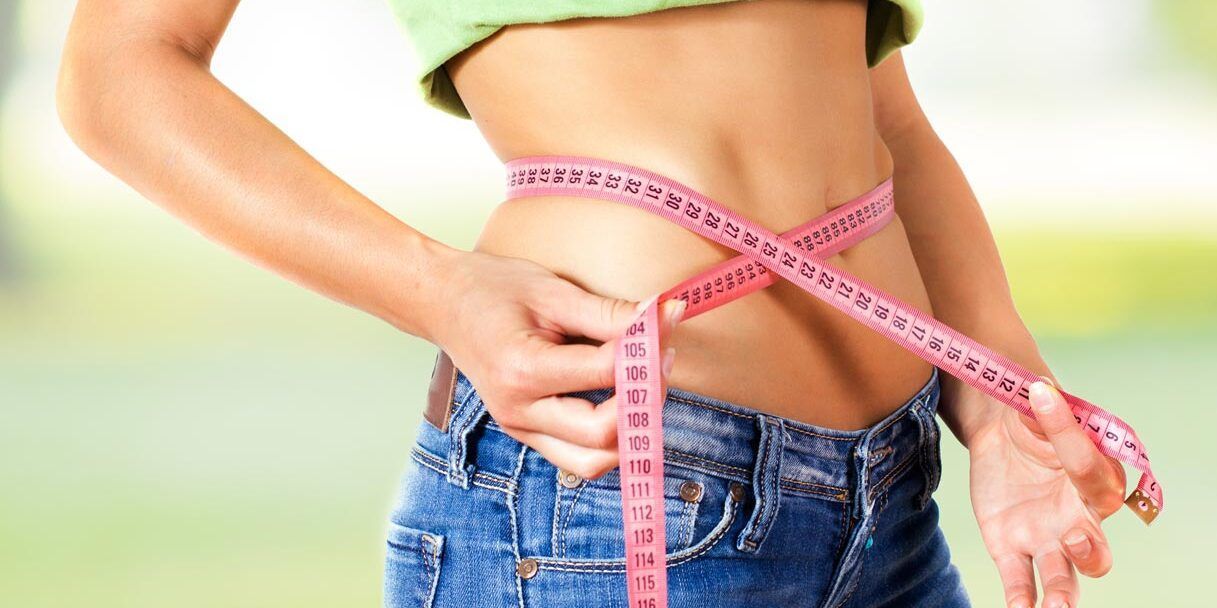 Donna che misura la vita con un metro a nastro rosa, indossa jeans e una maglietta verde.