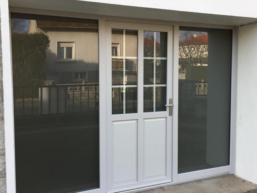 Porte blanche à panneaux de verre et imposte, encadrée de blanc sur un bâtiment blanc.