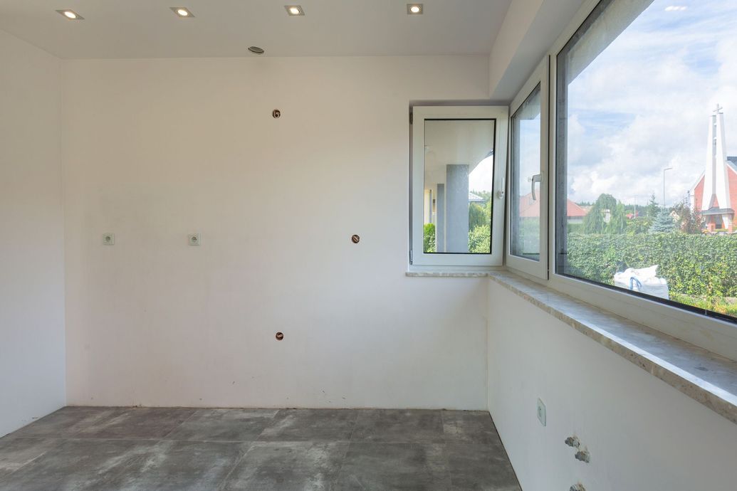 Pièce vide aux murs blancs, avec fenêtre et prises électriques apparentes ; sol gris.